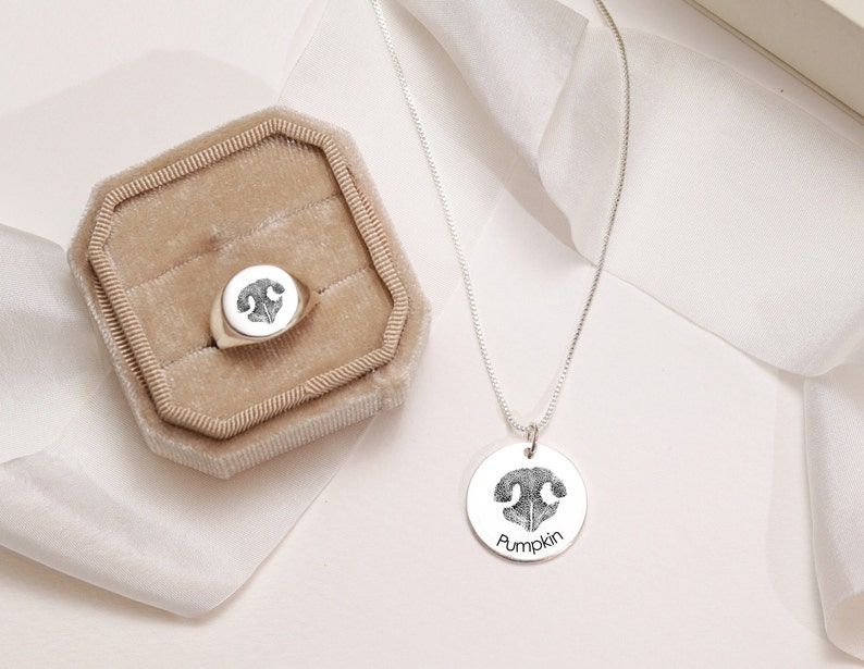 May include: A silver necklace with a round pendant featuring a black and white paw print. The pendant is engraved with the word "Pumpkin". A silver ring with a round top featuring a black and white paw print is in a brown velvet ring box.