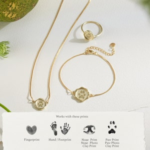 Peut inclure: Deux colliers en or avec un petit pendentif rond repr&eacute;sentant une empreinte de patte. Les colliers sont sur un fond blanc avec une carte qui dit "Fonctionne avec ces empreintes" et montre diff&eacute;rents types d'empreintes, y compris les empreintes digitales, les empreintes de mains/de pieds, les empreintes de nez et les empreintes de pattes.