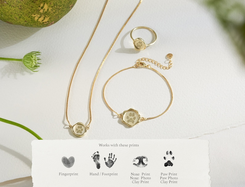 Peut inclure: Deux colliers en or avec un petit pendentif rond repr&eacute;sentant une empreinte de patte. Les colliers sont sur un fond blanc avec une carte qui dit "Fonctionne avec ces empreintes" et montre diff&eacute;rents types d'empreintes, y compris les empreintes digitales, les empreintes de mains/de pieds, les empreintes de nez et les empreintes de pattes.