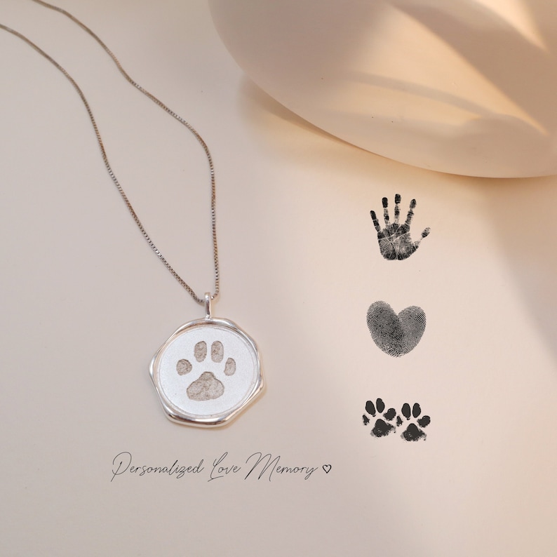 May include: A silver necklace with a round pendant featuring a paw print. The pendant is on a white surface with black ink paw prints, a heart, and a handprint.
