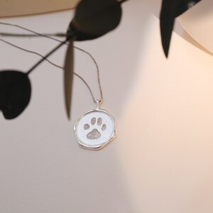 May include: A silver necklace with a round pendant featuring a paw print. The pendant is engraved with a detailed paw print.