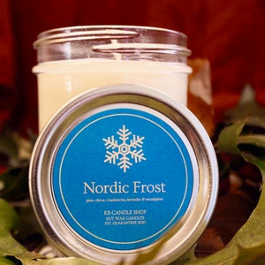 May include: A silver lid with a blue label that says "Nordic Frost" with a snowflake design. The label also says "pine, citrus, cranberry, lavender & eucalyptus" and "KB Candle Shop Soy Wax Candles Est. Quarantine 2020".