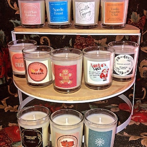Puede incluir: Una colección de 10 pequeños frascos de vidrio para velas con varios aromas navideños. Las velas están dispuestas en dos niveles de un soporte blanco de varios niveles. Las velas tienen etiquetas con diseños y textos festivos, que incluyen "Nordic Frost", "Vanilla Pumpkin Chai", "Santa's Sack", "Winter Woods", "Caramelized Pralines" y "Cinnamon Stick".