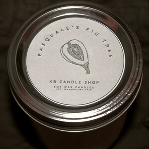 May include: A silver jar with a white label that reads "Pasquale's Fig Tree" and "KB Candle Shop Soy Wax Candles Est. Quarantine 2020". The label also features a black and white illustration of a fig.
