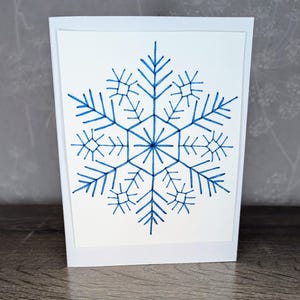 Può includere: Un biglietto bianco con un disegno di fiocco di neve blu. Il fiocco di neve è disegnato in uno stile geometrico semplice.