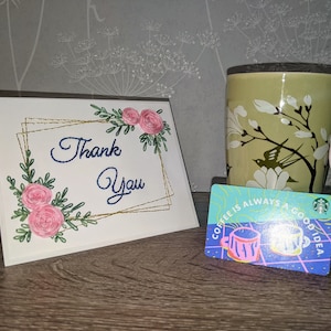 May include: A white card with gold embroidery of flowers and the text "Thank You". A green mug with white flowers and a bird design. A blue and green gift card with the text "Coffee is always a good idea" and a cartoon image of two coffee mugs.
