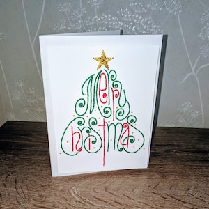May include: A white Christmas card featuring an embroidered Christmas tree design in green and red. The words "Merry Christmas" are stitched in a decorative style, topped with a gold star. The card is presented on a wooden surface.