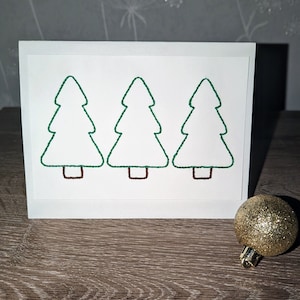 May include: A white card with three green embroidered Christmas trees. The trees are outlined in brown thread.