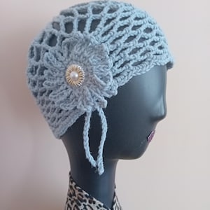 May include: A pale blue crocheted hat featuring a floral decoration with a pearl button. The hat has a net-like pattern and side ties. The hat is displayed on a black mannequin head.