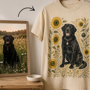 May include: A cream-colored short-sleeve t-shirt featuring a graphic illustration of a black Labrador retriever sitting among yellow sunflowers and small wildflowers. The dog is depicted with a collar and tag, matching the photo in the frame.
