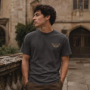 May include: A charcoal gray short-sleeve t-shirt featuring a small, detailed graphic of a moth printed on the left chest area. The shirt has a crew neckline and a standard fit, constructed from a solid-colored fabric with a smooth texture.