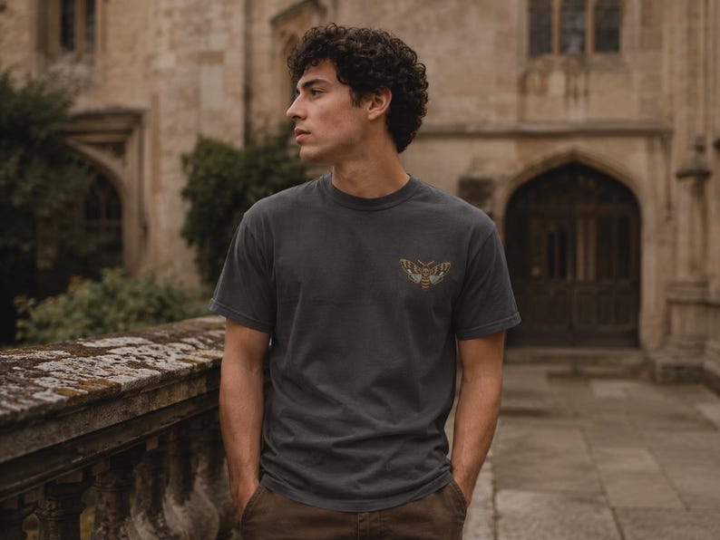 May include: A charcoal gray short-sleeve t-shirt featuring a small, detailed graphic of a moth printed on the left chest area. The shirt has a crew neckline and a standard fit, constructed from a solid-colored fabric with a smooth texture.