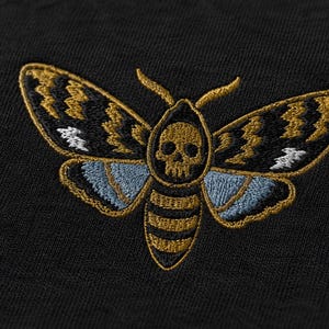 May include: A black fabric garment featuring an embroidered moth design. The moth has a skull pattern on its thorax, with wings detailed in gold, black, and light blue thread. The embroidery is centered on the dark, textured textile surface.