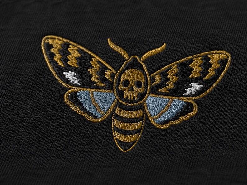 May include: A black fabric garment featuring an embroidered moth design. The moth has a skull pattern on its thorax, with wings detailed in gold, black, and light blue thread. The embroidery is centered on the dark, textured textile surface.