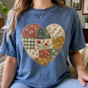 May include: A blue short-sleeved t-shirt featuring a large heart-shaped graphic on the chest. The heart is composed of various patchwork-style fabric patterns, including floral prints, small white flowers, and a green and white gingham check design.