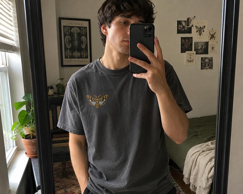 May include: A charcoal gray short-sleeve t-shirt featuring a small, detailed graphic of a moth with yellow, black, and white wings on the upper left chest. The shirt has a crew neckline and a relaxed fit, showcasing a muted, washed-out fabric texture.