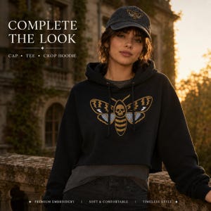 May include: A black cropped hoodie featuring a large embroidered moth graphic with a skull on its thorax, paired with a matching black baseball cap that has a smaller embroidered moth design on the front panel, both in a dark, distressed fabric.