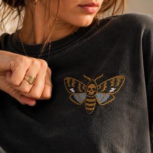 May include: A charcoal grey t-shirt featuring an embroidered moth design on the chest. The moth has a skull pattern on its thorax, with wings detailed in gold, light blue, and white thread. The shirt has a crew neckline and short sleeves.