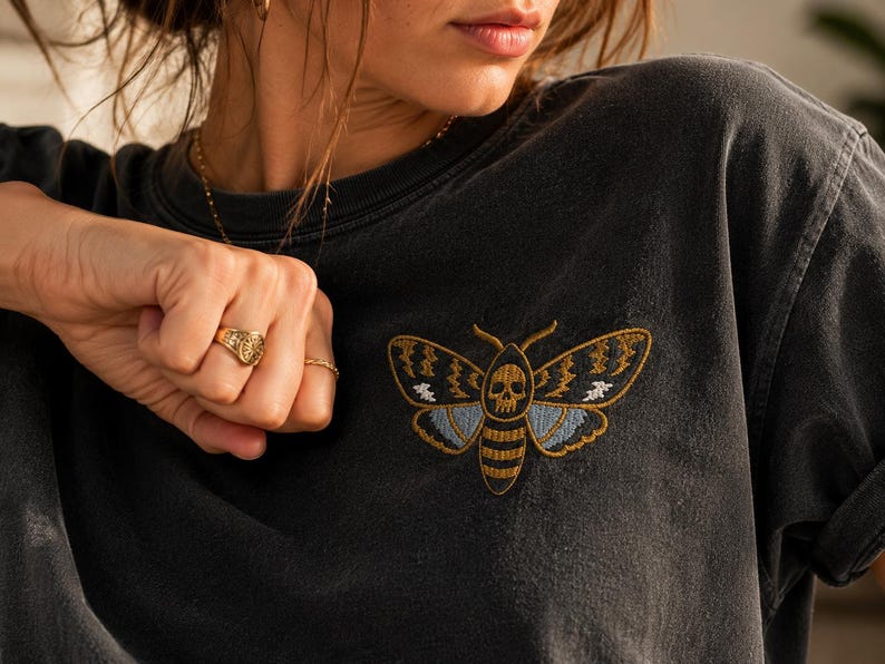May include: A charcoal grey t-shirt featuring an embroidered moth design on the chest. The moth has a skull pattern on its thorax, with wings detailed in gold, light blue, and white thread. The shirt has a crew neckline and short sleeves.