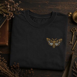 May include: A black crew neck t-shirt featuring an embroidered design on the chest. The embroidery depicts a moth with a skull pattern on its thorax, rendered in shades of gold, white, and grey, with detailed wing markings and a striped abdomen.