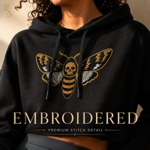 May include: A black hooded sweatshirt featuring an embroidered moth design on the chest. The moth has a skull pattern on its thorax, with wings detailed in gold, black, and light blue thread. The hoodie includes black drawstrings with metal tips.