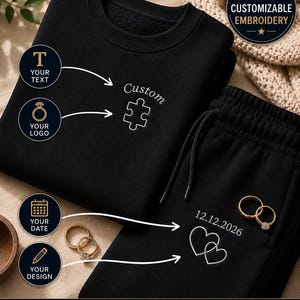 以下が含まれることがあります： カスタム刺繍オプション付きの黒いスウェットシャツとスウェットパンツのセット。スウェットシャツには「Custom」の文字とパズルのデザインが施されています。スウェットパンツには日付「12.12.2026」と2つのハートが表示されています。金の指輪も表示されています。