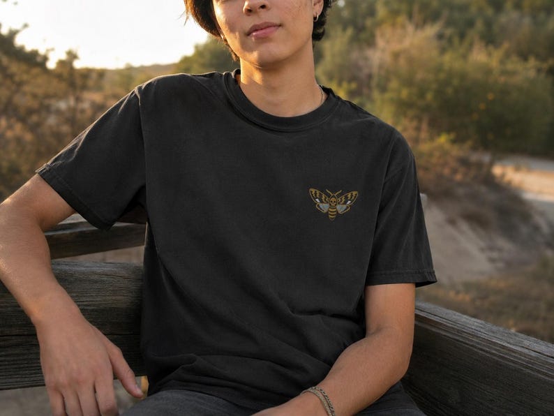 May include: A black short-sleeved t-shirt featuring a small, embroidered moth graphic on the left chest. The moth design is rendered in shades of yellow, brown, and white, showcasing detailed wing patterns and a central body structure.