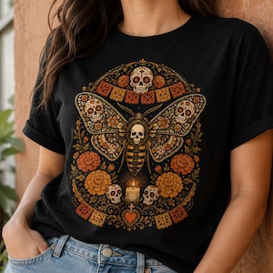 May include: A black t-shirt featuring a graphic print of a large moth with skull-patterned wings, surrounded by orange marigold flowers, small skulls, a lit candle, and decorative papel picado banners in a symmetrical, ornate arrangement.