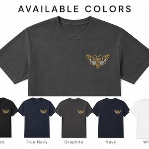 May include: A charcoal gray cropped t-shirt featuring a moth graphic with a skull pattern on the chest. Below, five t-shirts are displayed in black, true navy, graphite, navy, and white, each with the same moth graphic and color labels.