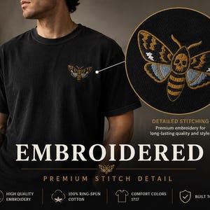 May include: A black t-shirt featuring a small embroidered moth design on the chest. The moth has a skull pattern on its thorax, with gold, blue, and white thread details. The image includes a circular inset showing a close-up of the embroidery.