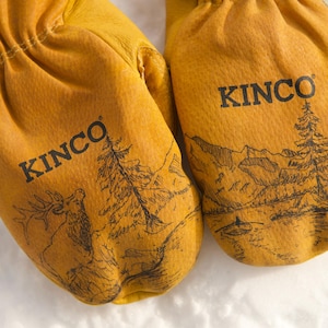 Puede incluir: Un par de manoplas de cuero amarillo con puños elásticos negros. Cada manopla presenta el texto negro KINCO y un dibujo detallado de líneas negras de un paisaje de montaña con árboles, un alce y un bote.
