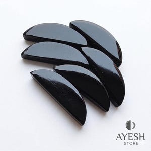 Puede incluir: Colección de siete piedras negras pulidas, semicirculares, dispuestas sobre una superficie blanca. Las piedras tienen un acabado brillante y una textura suave. La imagen incluye el logotipo "AYESH STORE" en la esquina inferior derecha.