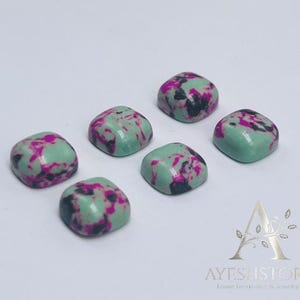 May include: Six square-shaped gemstones with a polished finish. The stones have a light green base color with a pattern of dark pink and black spots. The gemstones are arranged on a white surface.