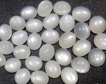 Puede incluir: Un conjunto de cabujones de piedra lunar blancos de forma ovalada. Las piedras tienen un brillo nacarado y están dispuestas juntas sobre una superficie oscura y texturizada. Las piedras preciosas son lisas y pulidas, reflejando la luz.