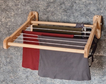 May include: A wooden wall-mounted clothes drying rack with four lines of clothesline. The rack has a natural wood finish and is folded up against the wall. Three pieces of clothing are hanging on the rack, including a red shirt, a gray shirt, and a green shirt.