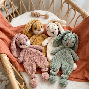 May include: Four plush crochet bunny loveys in dusty pink, mustard yellow, cream, and sage green. Each bunny features long floppy ears, a soft textured body, and knotted feet. Two bunnies have open eyes, while two have closed eyes with stitched details.