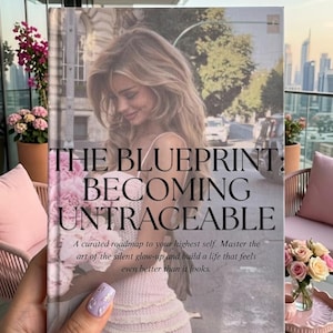 Könnte beinhalten: Ein Buch mit dem Titel "THE BLUEPRINT: BECOMING UNTRACEABLE" wird hochgehalten und zeigt das Bild einer Frau. Der Text auf dem Cover verspricht einen Fahrplan zur Selbstverbesserung und ein besseres Leben. Das Buch wird vor einem Balkon mit Stadtblick gehalten.