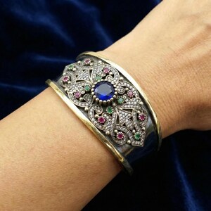 Könnte beinhalten: Ein silbernes Armreif-Armband mit goldfarbenem Besatz und aufwendigem Filigran-Design. Das Armband hat einen großen blauen Edelstein in der Mitte, umgeben von kleineren roten, grünen und klaren Edelsteinen. Das Armband befindet sich an einem Handgelenk.
