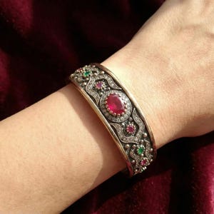 Op de afbeelding: Zilverkleurige manchetarmband met een ovale rode edelsteen in het midden, omringd door kleinere rode en groene edelstenen. De armband heeft een sierlijk ontwerp met ingewikkelde details.