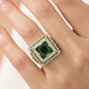 May include: A gold-toned ring with a large, square, emerald-green gemstone at its center. The ring is further embellished with smaller green and white gemstones, creating a striking, geometric design. The ring is a statement piece of jewelry.