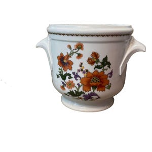 May include: A white ceramic planter with two side handles and a pedestal base. The exterior features a colorful floral pattern with orange, green, and purple flowers and leaves, accented by a thin brown decorative border along the top rim.