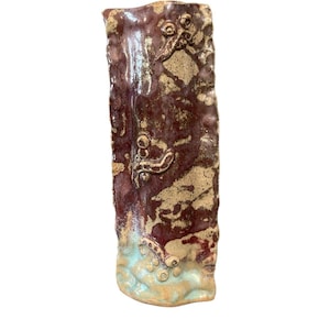 May include: A handmade, rectangular ceramic vase with a textured surface. The vase features a deep burgundy color with tan and light blue accents. Decorative raised elements are visible on the surface, adding to its unique, artistic appeal.
