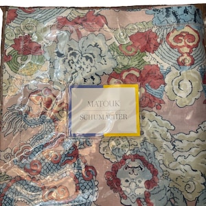 May include: A folded fabric textile featuring a multi-colored pattern of dragons and clouds in shades of pink, blue, red, and cream. A rectangular label is attached to the center, displaying the text MATOUK SCHUMACHER in dark gray lettering.