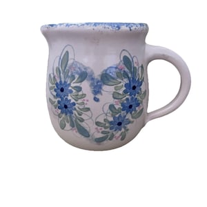 May include: White ceramic mug with a rounded body and a handle. The mug is decorated with blue floral designs and green leaves. The rim has a blue speckled pattern. A charming piece for enjoying your favorite beverage.