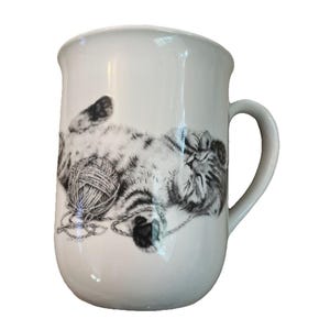 May include: A white ceramic mug featuring a detailed black and white sketch of a fluffy kitten lying on its back. The kitten is depicted playing with a ball of yarn, with its paws tangled in the string. The mug has a smooth, rounded handle.