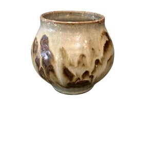 May include: A small, round ceramic vase with a wide opening. The vase has a light beige base colour with brown, organic-shaped patterns. The rim is a darker brown. The overall shape is bulbous, with a slightly uneven texture.