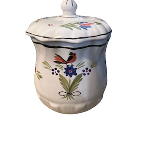 May include: White ceramic jar with a lid, adorned with hand-painted floral and bird motifs. The jar features a black rim and a knob on the lid. The design includes red, blue, and green accents, creating a charming, decorative piece.