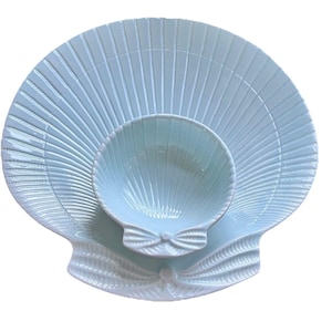 May include: A light blue ceramic serving dish shaped like a seashell with a smaller matching bowl inside. The dish has a ribbed texture and a raised bow detail.