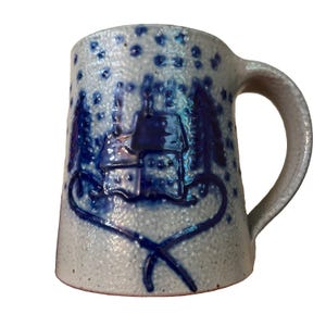 May include: A light gray speckled ceramic mug featuring a dark blue embossed design of a small house surrounded by pine trees and falling snow. The base of the house is framed by a dark blue line that curves into a heart shape at the bottom.