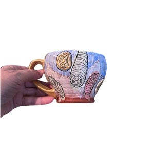 May include: A ceramic mug with a gold-colored handle featuring a hand-painted design of abstract, swirling oval shapes in shades of yellow, pink, and white against a light blue and textured pink background with a solid reddish-brown base.